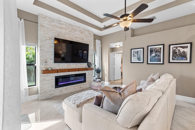 living room featuring ceiling fan, a tray ceiling, a stone fireplace, and baseboards