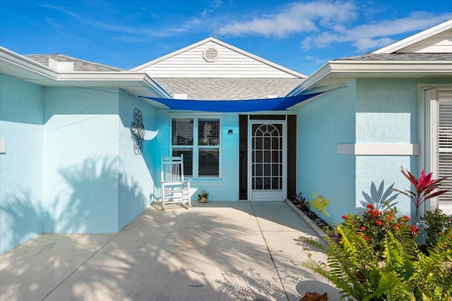 entrance to property featuring roof with shingles, a patio, and stucco siding