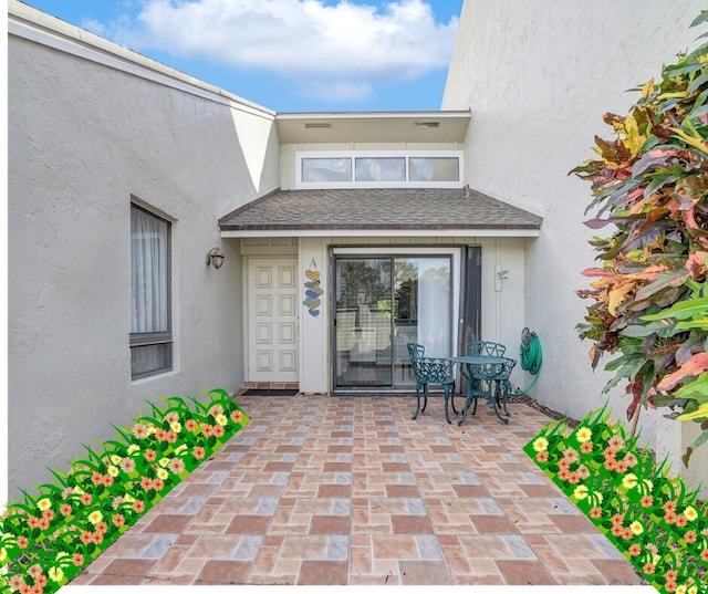 view of exterior entry featuring a patio area, roof with shingles, and stucco siding