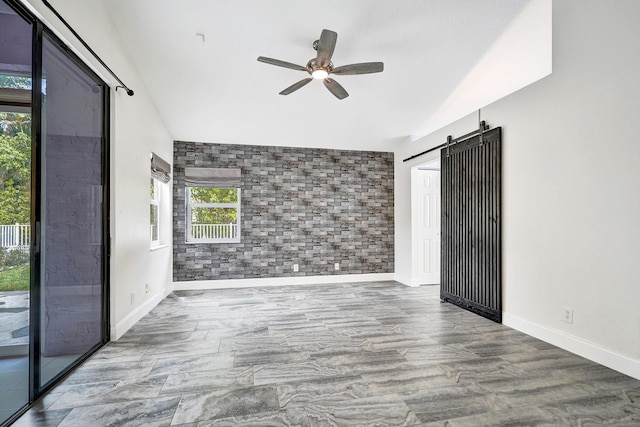 empty room with a barn door, an accent wall, brick wall, a ceiling fan, and baseboards
