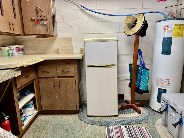 kitchen featuring electric water heater, light countertops, freestanding refrigerator, brown cabinets, and concrete block wall