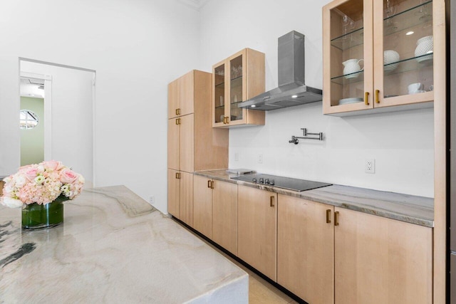 kitchen featuring wall chimney exhaust hood, glass insert cabinets, light stone counters, black electric stovetop, and light brown cabinets