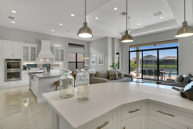 kitchen with custom range hood, visible vents, stainless steel double oven, open floor plan, and an island with sink