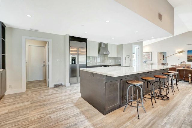 kitchen featuring modern cabinets, light stone counters, stainless steel built in refrigerator, a large island with sink, and wall chimney range hood