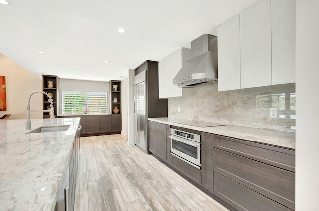 kitchen with light stone counters, a sink, wall chimney range hood, appliances with stainless steel finishes, and modern cabinets