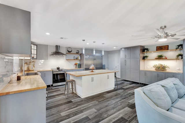 kitchen featuring appliances with stainless steel finishes, a kitchen breakfast bar, wall chimney range hood, open shelves, and wooden counters