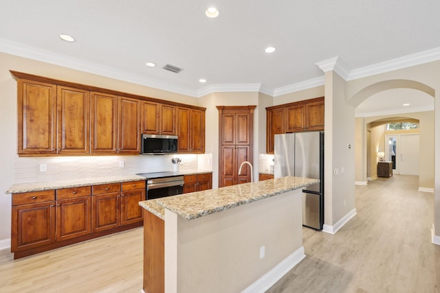 kitchen with arched walkways, appliances with stainless steel finishes, light stone countertops, light wood finished floors, and an island with sink