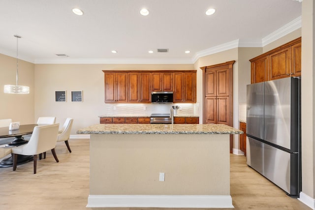 kitchen with a center island with sink, brown cabinetry, appliances with stainless steel finishes, hanging light fixtures, and light stone countertops