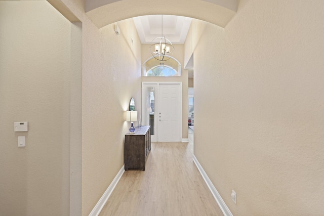 corridor featuring baseboards, arched walkways, an inviting chandelier, a tray ceiling, and light wood-style floors