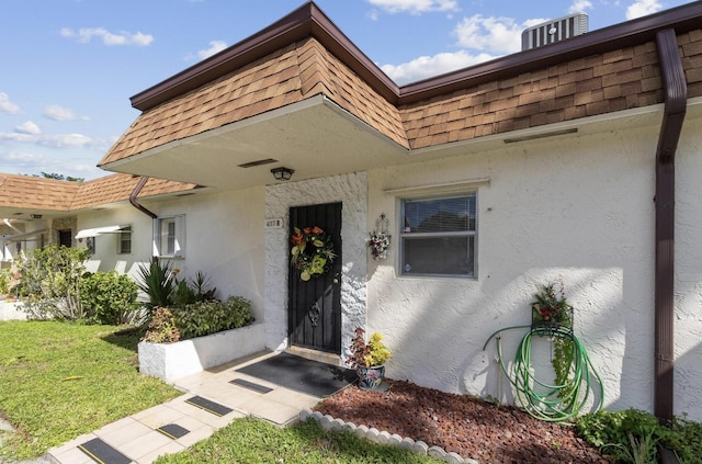 view of exterior entry featuring a yard, a shingled roof, mansard roof, and stucco siding