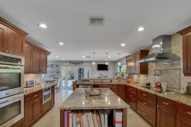 kitchen featuring visible vents, a peninsula, wall chimney range hood, double oven, and a sink