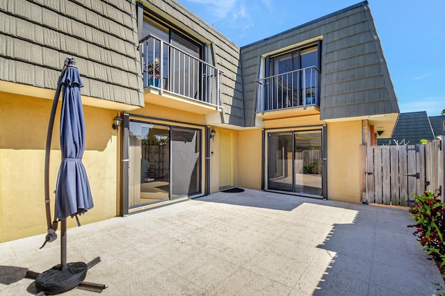 rear view of property featuring a patio, mansard roof, a balcony, fence, and stucco siding