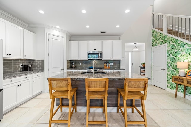 kitchen featuring stainless steel microwave, stone countertops, a center island with sink, and white cabinetry