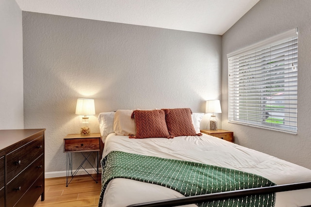 bedroom featuring light wood-type flooring, a textured wall, lofted ceiling, and baseboards