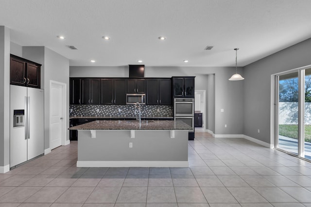 kitchen featuring a center island with sink, appliances with stainless steel finishes, light tile patterned flooring, dark stone countertops, and a kitchen breakfast bar