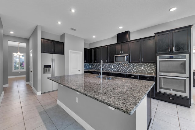 kitchen featuring visible vents, dark stone counters, an island with sink, stainless steel appliances, and a sink