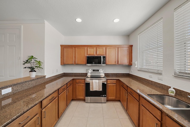 kitchen with light tile patterned flooring, a sink, appliances with stainless steel finishes, dark stone counters, and brown cabinetry