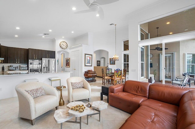 living room featuring light tile patterned floors, arched walkways, ornamental molding, a high ceiling, and ceiling fan with notable chandelier