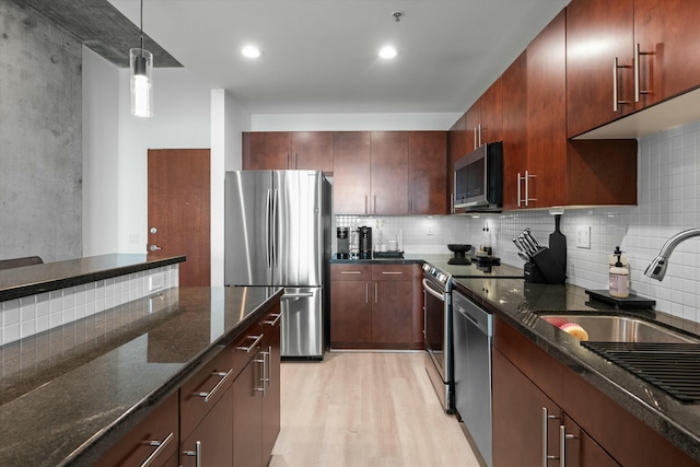 kitchen with a sink, appliances with stainless steel finishes, dark stone counters, light wood finished floors, and decorative light fixtures