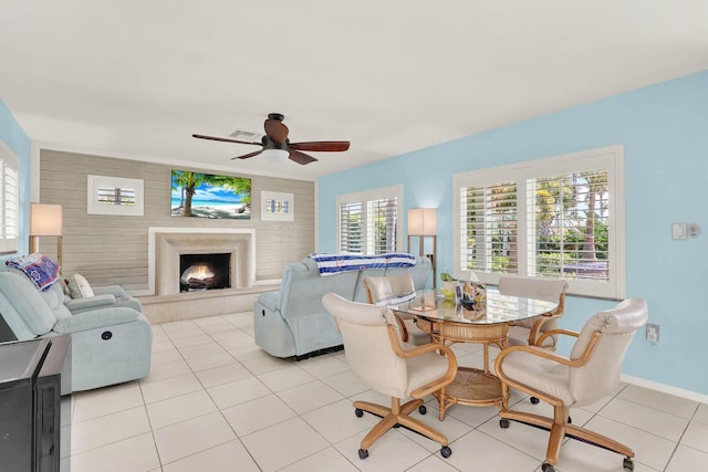 living room with light tile patterned floors, a warm lit fireplace, visible vents, baseboards, and ceiling fan