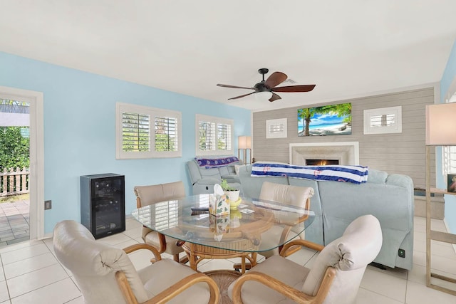 dining area with light tile patterned floors, a ceiling fan, wine cooler, an accent wall, and a lit fireplace