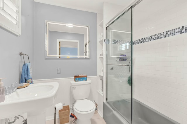 full bath with wainscoting, a sink, toilet, and bath / shower combo with glass door