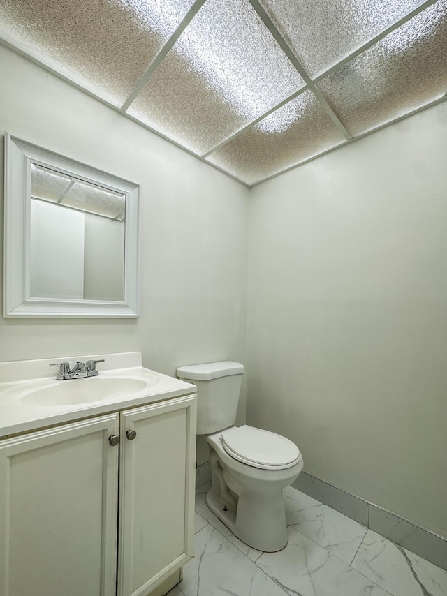 half bathroom featuring a paneled ceiling, toilet, vanity, baseboards, and marble finish floor