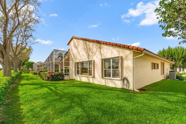 back of property featuring a lawn, glass enclosure, a tiled roof, central air condition unit, and stucco siding