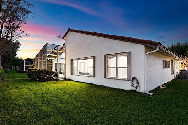 back of property at dusk with a yard, central AC, a lanai, and stucco siding