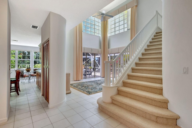 entrance foyer with light tile patterned floors, visible vents, a towering ceiling, a ceiling fan, and stairs