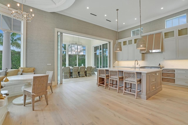 kitchen featuring light wood finished floors, light countertops, a sink, wall chimney range hood, and an island with sink