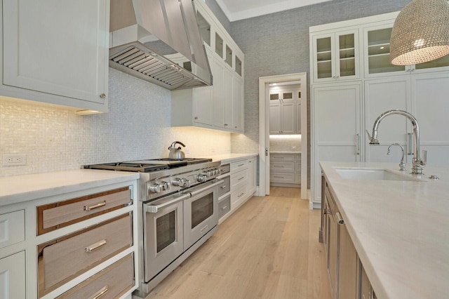 kitchen with range with two ovens, light wood-style flooring, a sink, wall chimney range hood, and tasteful backsplash