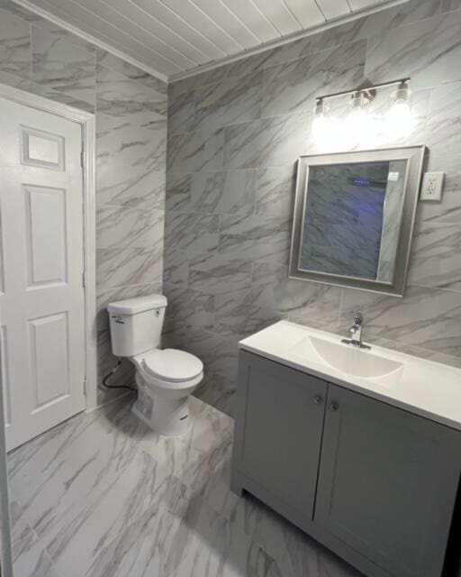 half bath featuring toilet, marble finish floor, vanity, and tile walls