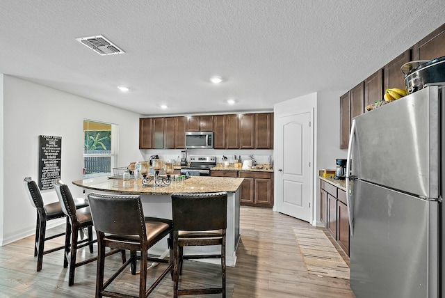 kitchen with dark brown cabinetry, a center island with sink, visible vents, appliances with stainless steel finishes, and light wood-type flooring