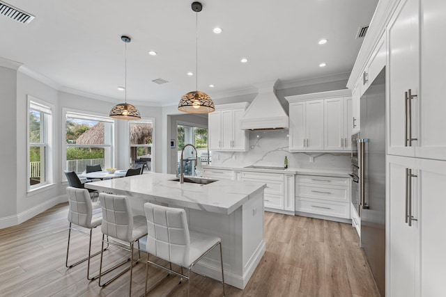 kitchen featuring visible vents, an island with sink, a sink, decorative backsplash, and custom range hood