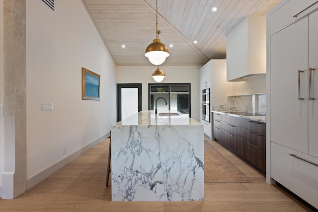 kitchen featuring dark brown cabinetry, white cabinets, an island with sink, wooden ceiling, and light stone counters