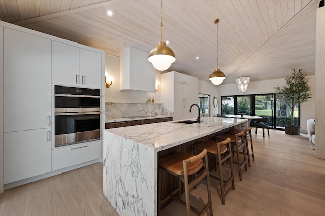 kitchen with light stone counters, decorative light fixtures, double oven, white cabinetry, and an island with sink