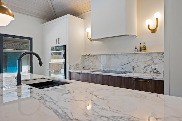 kitchen with black electric stovetop, stainless steel double oven, a sink, white cabinets, and wall chimney range hood