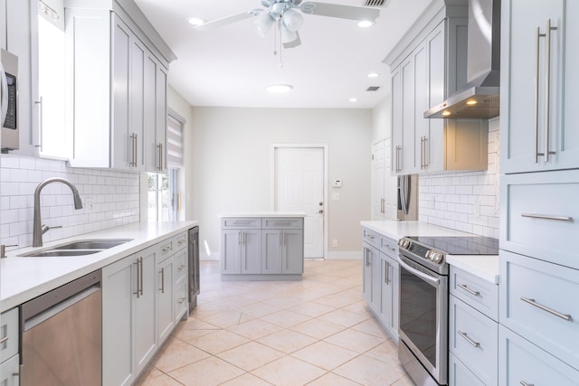 kitchen with gray cabinetry, stainless steel appliances, a sink, light countertops, and wall chimney range hood