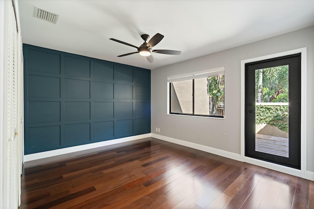 empty room with dark wood-style floors, visible vents, baseboards, ceiling fan, and a decorative wall