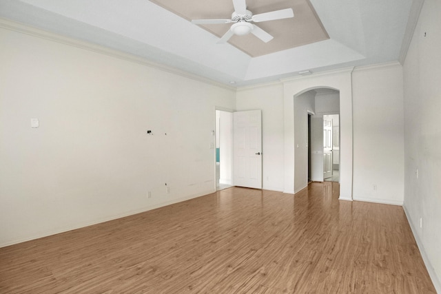 empty room with arched walkways, ceiling fan, wood finished floors, a tray ceiling, and crown molding