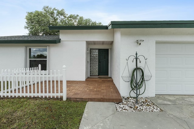 view of exterior entry featuring stucco siding, a garage, a shingled roof, and fence