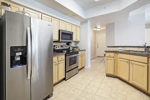 kitchen featuring light tile patterned floors, stainless steel appliances, a sink, light brown cabinetry, and dark stone countertops