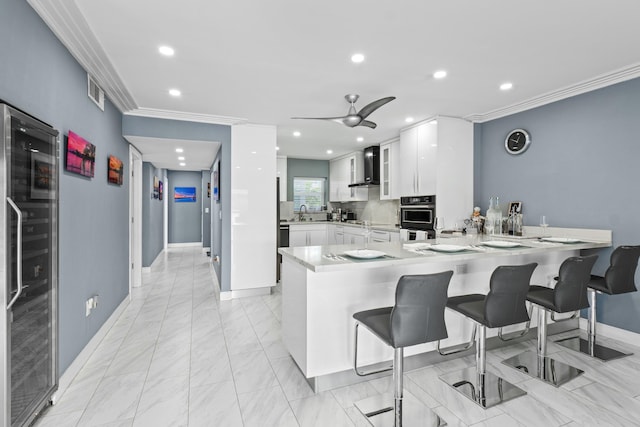 kitchen with wall chimney exhaust hood, glass insert cabinets, ornamental molding, a peninsula, and white cabinetry