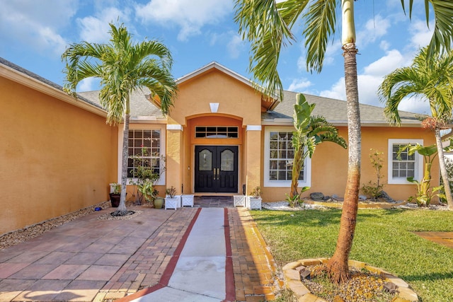 view of exterior entry featuring stucco siding, french doors, a lawn, and roof with shingles