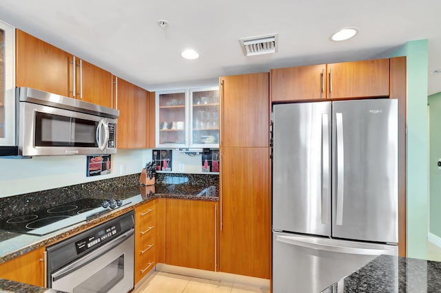 kitchen featuring brown cabinets, stainless steel appliances, visible vents, glass insert cabinets, and dark stone countertops