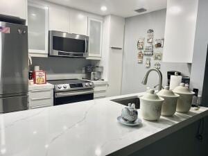 kitchen with stainless steel appliances, a sink, white cabinetry, light stone countertops, and glass insert cabinets