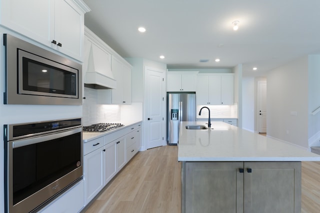 kitchen with a center island with sink, white cabinets, appliances with stainless steel finishes, light wood-style floors, and premium range hood