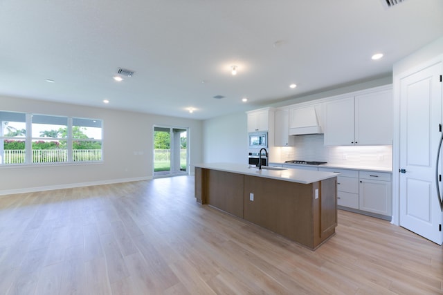 kitchen with a center island with sink, black cooktop, visible vents, stainless steel microwave, and premium range hood