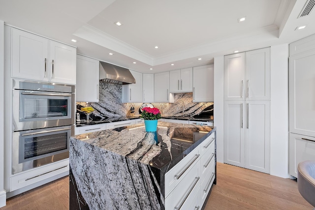 kitchen with light wood-type flooring, wall chimney range hood, a tray ceiling, and stainless steel double oven